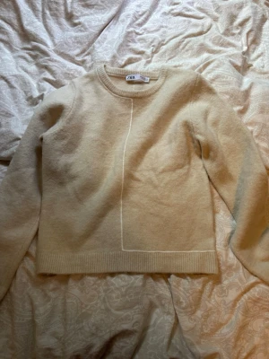 Beige tröja från Zara - Mysig beige tröja från Zara. Tröjan har rund halsringning, långa ärmar och ribbade muddar. En diskret vit linje går vertikalt och horisontellt på framsidan för en stilren look.