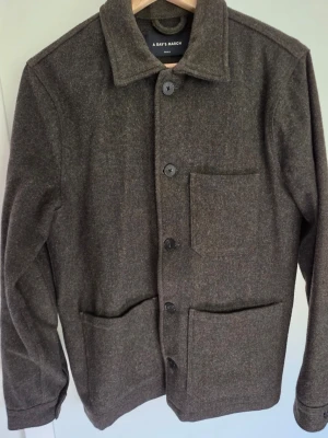 Mörkgrön overshirt från A Day's March - Inga defekter. Stilren mörkgrön overshirt från A Day's March i ullmix. Klassisk krage, stora framfickor och knäppning med svarta knappar. Perfekt lager-på-lager-plagg för kyligare dagar och enkel att matcha med olika outfits. 