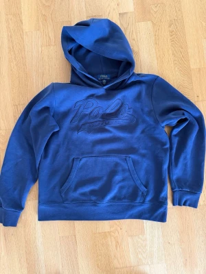 Blå hoodie Polo Ralph Lauren, Junior L - Snygg blå hoodie från Polo Ralph Lauren med stor broderad logga på bröstet. Klassisk känguruficka framtill och mjukt material som känns skönt mot huden. Perfekt för dig som vill ha en stilren och bekväm hoodie med streetkänsla. Junior large (14-16)