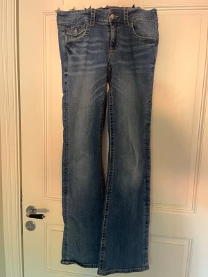 Blå low waist bootcut jeans med fickdetalj - Säljer ett par blå bootcut jeans med snygga slitningar och kontrastsömmar. Jeansen har en unik fickdetalj med lock och knapp framtill samt klassisk femficksmodell. Jeansen är också lågmidjade och tillverkade i denim med lätt stretch för skön passform. Perfekta till sneakers eller boots.