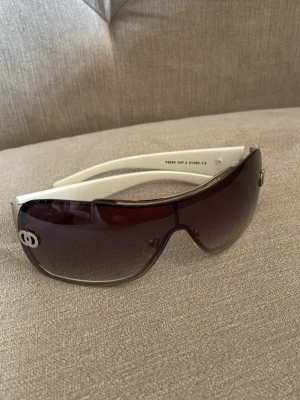 Vita solglasögon med stora bågar - Säljer ett par snygga solglasögon med vita skalmar och stora, mörka glas i shield-modell. Glasen har en lätt toning och bågarna har en silverfärgad detalj på sidan. Perfekta för dig som vill sticka ut i sommar!