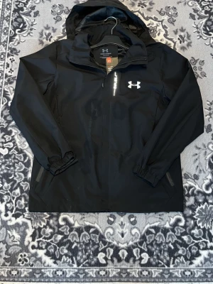 Svart regnjacka från Under Armour - Snygg svart regnjacka från Under Armour med huva och dragkedja framtill. Jackan har elastiska muddar vid ärmsluten, två sidofickor med dragkedja och logga på bröstet. Tillverkad i vattenavvisande material, perfekt för regniga dagar.