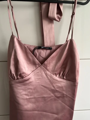 Rosa top med smala band - Snygg rosa top  i satinliknande material med smala axelband och v-ringning. Klänningen har knytband i midjan och en elegant, glansig finish som ger en lyxig känsla. Perfekt för dig som vill ha en feminin och trendig look.