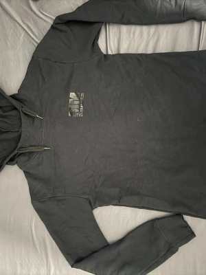 Svart hoodie med tryck från MM SPORTS - Cool svart hoodie med huva och snörning. Framtill finns ett diskret tryck och på ryggen står det 'NN SPORTS' i stort, slitet tryck. Perfekt för en avslappnad streetstyle-look. Mjuk bomullskänsla och klassisk passform.
