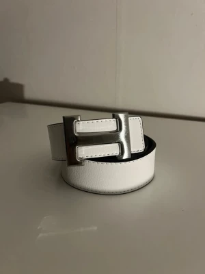 Vitt brett bälte med silverfärgat spänne - Snyggt vitt bälte i skinnimitation med ett brett, rektangulärt silverfärgat spänne. Bältet har en stilren och modern look som passar till jeans eller byxor. Perfekt accessoar för att lyfta din outfit.
