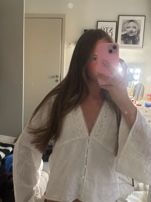 Vit broderad blus med vida ärmar - Säljer en vit blus med broderade blommor och v-ringning. Blusen har vida långärmade ärmar och knappar framtill. Detaljerad spetskant vid halsen ger en romantisk känsla. Perfekt för dig som gillar boho-stil och lätta material.