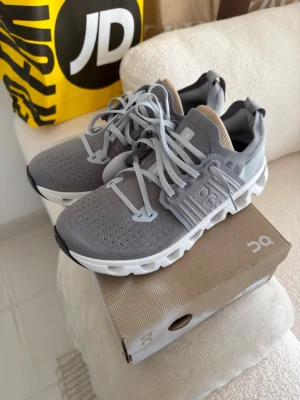 Grå On Cloud sneakers - Stilrena grå On Cloud sneakers med vit sula och ljusgrå detaljer. Ovandelen är i ventilerande mesh och skorna har en unik, chunky sula för extra komfort. Klassisk snörning och diskret logga på sidan. Perfekta för dig som gillar sportig och modern stil.