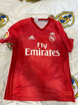 Real Madrid tröja - Snygg röd Real Madrid matchtröja från Adidas med vitt tryck, klubbmärke och Fly Emirates-logga på bröstet. La Liga-märke på ärmen och klassisk V-ringning. Tillverkad i lätt och ventilerande material, perfekt för fotboll eller träning. Som på bilden så är trycket på ryggen borta och laliga märket slitet. 