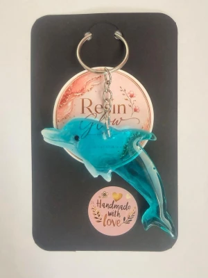 Nyckelring med delfin i resin - Säljer en handgjord nyckelring med en delfin i transparent blå resin. Delfinen har fina detaljer och sitter fast i en metallkedja med ring. Perfekt att hänga på väskan eller nyckelknippan för en unik touch.