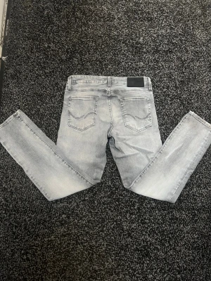 Slim/Glenn jack & jones jeans - Säljer ett par ljusgrå jeans från Jack & Jones med klassisk femficksdesign och snygga sömdetaljer på bakfickorna. Modellen är slim/glenn