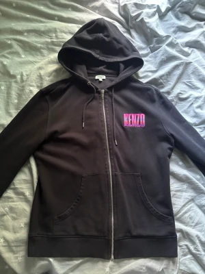 Svart Kenzo hoodie med neontryck - Säljer en svart hoodie från Kenzo med dragkedja och huva. På bröstet finns ett rosa-blått Kenzo-tryck och på ryggen ett stort neonfärgat Kenzo-motiv med berg och sol. Hoodien har fickor fram och ribbade muddar. Perfekt för dig som gillar streetwear och snygga detaljer.