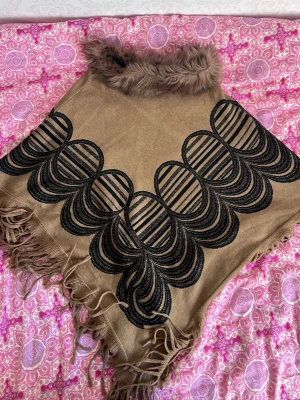 Brun poncho med fuskpäls och fransar - Snygg brun sjal/poncho med mjuk fuskpäls vid halsen och dekorativa svarta broderier. Sjalen har långa fransar längs kanterna och är tillverkad i ett mjukt tyg som känns skönt mot huden. Perfekt för att lyfta din outfit med en unik touch. Endast använd 1-3 gånger, helt ny mycket god skick😊