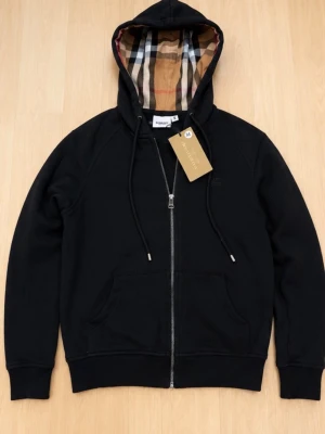 Svart hoodie från Burberry med dragkedja - Svart hoodie från Burberry i storlek M med dragkedja framtill och klassiskt rutigt foder i huvan. Hoodien har dragsko i huvan, två fickor fram och ribbade muddar. Perfekt för en stilren och lyxig look.