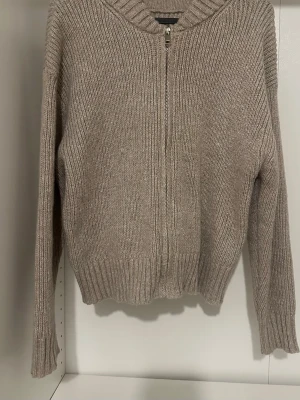 Beige stickad ziptröja - Mysig beige stickad tröja med dragkedja framtill och ribbade muddar vid ärmslut och nederkant. Tröjan har en avslappnad passform och är perfekt för lager på lager. Materialet ser ut att vara en mjuk ull- eller ullmix.