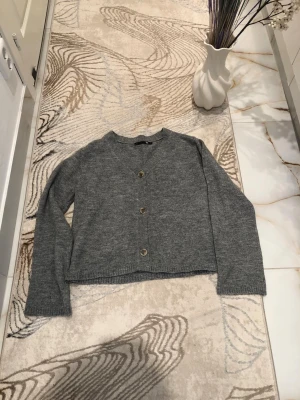 Grå basic kofta med knappar - En enkel och stilren grå kofta med tre knappar framtill. Koftan har v-ringning, långa ärmar och är gjord i mjukt stickat material. Perfekt att slänga över en t-shirt för en avslappnad look.