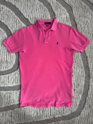 Rosa pikétröja från Polo Ralph Lauren - Klassisk rosa pikétröja från Polo Ralph Lauren med blå broderad logga på bröstet. Tröjan har korta ärmar, krage och knappar framtill. Tillverkad i bomull för en bekväm och luftig känsla.