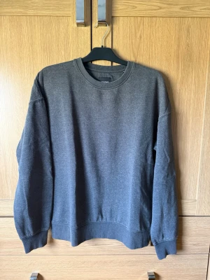 Mörkgrå sweatshirt från Jack & Jones - En stilren mörkgrå sweatshirt från Jack & Jones med rund halsringning och ribbade muddar vid ärmslut och nederkant. Tröjan har en klassisk passform och är tillverkad i mjukt bomullsmaterial. Perfekt för en avslappnad look.