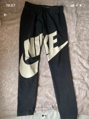Svarta leggings från Nike med logga - Snygga svarta leggings från Nike med stor vit logga och swoosh på ena benet. Resår i midjan och texten 'JUST DO IT.' på insidan. Perfekta för en sportig och avslappnad stil. Tillverkade i mjukt och stretchigt material för bästa komfort.