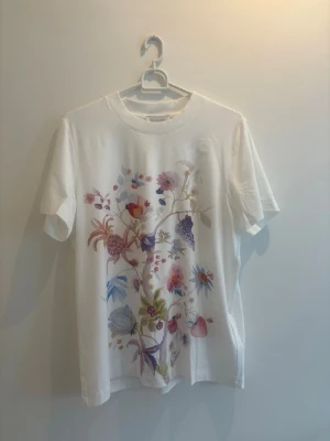 Vit t-shirt med blommigt tryck - Snygg vit t-shirt med färgglatt blommigt tryck på framsidan. T-shirten har rund hals och korta ärmar, perfekt för dig som gillar en fräsch och stilren look med en twist. Materialet känns mjukt och skönt mot huden.