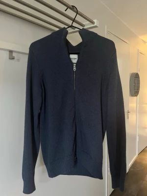 Mörkblå ziptröja från Dobber - Mörkblå stickad ziptröja från Dobber med hög krage och hel dragkedja framtill. Tröjan har ribbade muddar vid ärmslut och nederkant, vilket ger en snygg och bekväm passform. Perfekt för lager på lager och enkel att matcha.