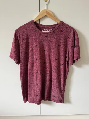 Vinröd t-shirt med småmönster Knickerbocker Vintage strl S Design strl  - Säljer en vinröd t-shirt med små svarta mönster över hela plagget. T-shirten har rund halsringning och korta ärmar. Från knickerbocker vintage design. Material 60% bomull, 40% polyester. Storlek S.