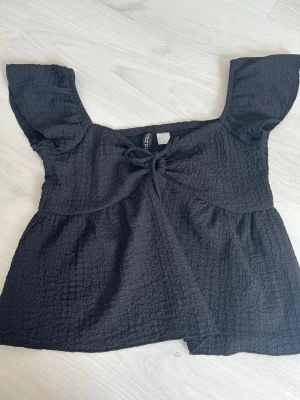 Svart topp med knyt och puffärm - Säljer en svart topp med puffiga korta ärmar och knyt framtill. Toppen har en strukturerad, lite krinklad yta och en luftig passform. Perfekt till jeans eller kjol för en trendig look.