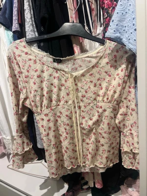 Blommig topp - Söt blus i cremevitt med små rosa blommor och volangdetaljer på ärmarna. Blusen har lång ärm, knytband framtill och lätt rynkad struktur. Materialet är stretchigt och mjukt, perfekt för en romantisk look. Står storlek M men är passar bättre för S