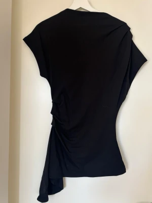 Svart asymmetrisk klänning - Svart topp med asymmetrisk design och draperad detalj på sidan. Toppen har korta ärmar och ett långt tygstycke som hänger ner på ena sidan för en unik look. Tillverkad i ett mjukt material som ger en snygg passform.