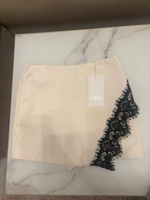 Krämvit byx/kjol med spets från ZARA - Supersnygg krämvit skort från ZARA med svart spetsdetalj längs ena sidan. Skorten har en rak passform och dragkedja bak. Materialet ser ut att vara mjukt och lite glansigt, perfekt för en trendig och chic look.