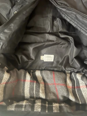 Svart pufferjacka från Burberry - Snygg svart pufferjacka från Burberry London England med klassiskt rutigt foder i grått, svart, vitt och rött. Jackan har en modern look och är tillverkad i ett glansigt material som ger en exklusiv känsla. Perfekt för kalla dagar när du vill hålla stilen. Den har hål på ärmarna som lätt kan sys ihop men de är därför priset är så billigt 