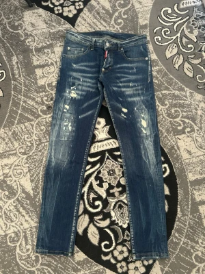 Dsquared2 Jeans - Säljer ett par blå jeans med slitningar och distressed detaljer framtill. Jeansen har raka ben, klassisk femficksmodell och en patch baktill i midjan. Perfekta för en avslappnad och trendig look.
