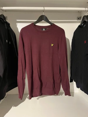 Vinröd stickad tröja Lyle & Scott - Vinröd stickad tröja med rund hals och lång ärm. Tröjan har en gul broderad logga på bröstet och ribbade muddar vid ärmslut och nederkant. Tröjan är L men har krympt till storlek S. Perfekt för dig som gillar stilrena och klassiska plagg.