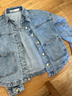 Ljusblå oversized jeansjacka från Zara - Snygg ljusblå oversized jeansjacka från Zara med stora bröstfickor och guldfärgade knappar. Klassisk krage och avslappnad passform som ger en cool och trendig look. Perfekt att slänga över en hoodie eller t-shirt för en casual vibe.