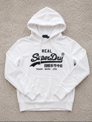 Vit hoodie från Superdry med tryck - Säljer en vit hoodie från Superdry med svart logga och japansk text på bröstet. Klassisk känguruficka framtill, ribbade muddar och dragsko i huvan. Perfekt för dig som gillar streetwear och vill ha en stilren look.