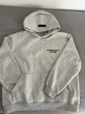  Essentials  hoodie - Säljer en grå hoodie från Essentials Fear of God med svart logga på bröstet. Hoodien har en stor huva, ribbade muddar och magficka. Tillverkad i mjukt bomullsmaterial som är perfekt för chill dagar. Enkel och clean design som passar till allt.