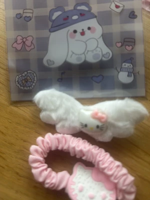 Sött Hello Kitty håraccessoarset - Tvådelat håraccessoarset med en fluffig vit hårklämma med Hello Kitty och vingar samt en rosa scrunchie med en söt Hello Kitty-detalj. Perfekt för dig som gillar gulliga och lekfulla detaljer i håret.