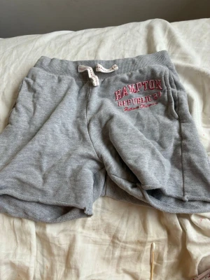 Grå mjukisshorts med snörning - Grå mjukisshorts från Hampton Republic 27 med vit snörning i midjan och tryckt logga i rosa på vänster sida. Sköna shorts i bomullsmix, perfekta för chill dagar eller träning. Resår i midjan för extra komfort.