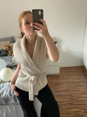 Beige stickad omlottkofta med knytband - Säljer en beige stickad omlottkofta med bred krage och knytband i midjan. Koftan är ärmlös och har en ribbad struktur som ger en mysig och trendig look. 