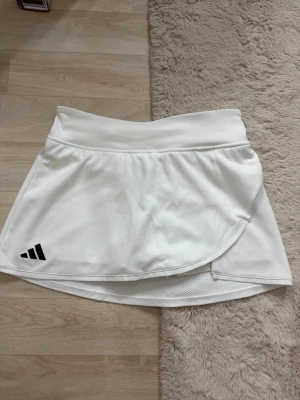 Vit sportig kjol från Adidas -  Snygg vit kjol från Adidas med bred midjeresår och diskret struktur i tyget. Kjoldesignen har inbyggda shorts under och en svart Adidas-logga framtill. Perfekt för en aktiv och trendig look.