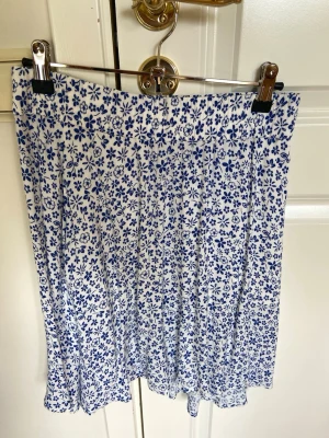En kjol i vit och blå från Bondelid i storlek XS - Söt vit kjol med blått blommönster. Kjolen har en lätt utställd form och är knälång, perfekt för varma dagar. Tillverkad i ett mjukt och skönt material som känns luftigt. Passar dig som gillar en fräsch och somrig stil.