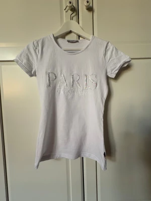 Vit t-shirt med PARIS-brodyr - Säljer en vit t-shirt med broderad text 'PARIS MAGNIFIQUE' på bröstet. Bokstäverna har ett blommigt mönster i flera färger. T-shirten har rund hals och korta ärmar, perfekt för en avslappnad och trendig look.