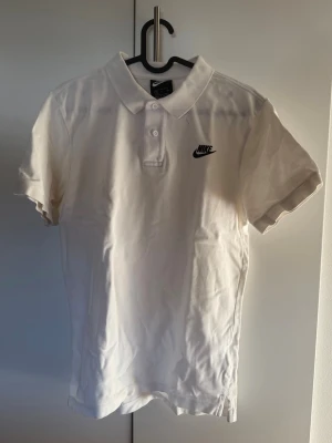 Vit pikétröja från Nike - Klassisk vit pikétröja från Nike med korta ärmar och krage. Tröjan har knappar framtill och en broderad Nike-logga på bröstet. Perfekt till en avslappnad och sportig stil.