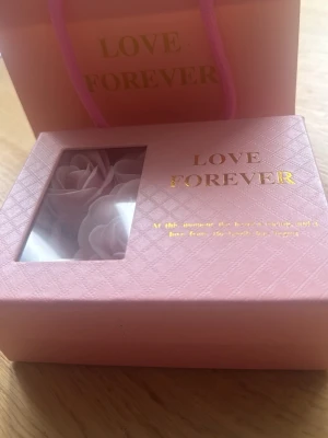 Presentask med rosa konstrosor - Söt presentask i ljusrosa med texten 'Love Forever' i guld. Innehåller fyra konstgjorda rosor i mjukt rosa färg, perfekta som dekoration eller gåva. Asken har ett fönster i locket och matchande presentpåse.