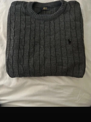 Grå kabelstickad tröja - Snygg grå kabelstickad tröja med rund halsringning och diskret broderad logga på bröstet. Tröjan har långa ärmar och ribbade muddar vid hals, ärmslut och nederkant. Perfekt för lager på lager och en klassisk look.