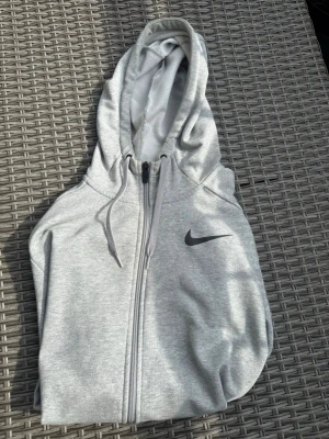 Ljusgrå zip hoodie från Nike - Snygg ljusgrå hoodie från Nike med dragkedja framtill och klassisk logga på bröstet. Tröjan har huva med dragsko och två fickor framtill. Tillverkad i mjukt sweatshirtmaterial som är skönt att ha på sig. Perfekt för en avslappnad och sportig stil. Endast testad så tröjan är i princip ny!