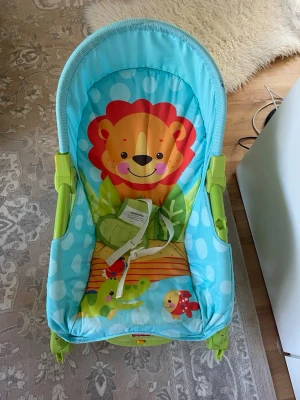Färgglad babysitter med lejonmotiv - Säljer en babysitter i blått och grönt med ett gulligt lejonmotiv och andra djur på tyget. Babysittern har säkerhetsbälte och robust plaststomme. Perfekt för små barn som vill sitta bekvämt och leka.