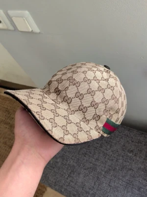 Beige Gucci keps (ÄKTA) - Säljer en beige keps från Gucci med det klassiska GG-monogrammet över hela kepsen. På sidan finns en grön och röd rand som ger extra lyxig känsla. Kepsen har böjd skärm och är tillverkad i canvasmaterial. Perfekt för dig som vill ha en stilren och trendig accessoar.