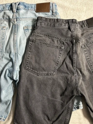 Två par jeansshorts i blått och grått - Säljer två par klassiska jeansshorts, ett par i ljusblå denim och ett par i mörkgrå denim. Båda har hög midja, raka ben och stora bakfickor. Perfekta för sommaren och enkla att matcha med olika stilar. Orginalpris - 500kr st