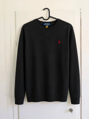 Polo Ralph Lauren sweatshirt svart 🖤❤️ - Klassisk svart sweatshirt från Polo Ralph Lauren med den ikoniska röda broderade hästloggan på bröstet. Stilren modell som passar till det mesta. • Storlek: L/G (14–16), ungdom men passar xs-s i vuxenstorlek!😁 • Färg: Svart med röd logga • Rund hals • Ribbstickade muddar och nederkant • Tillverkad i Kambodja • Fint skick utan defekter! Säljer då den inte kommer till användning längre. 📦