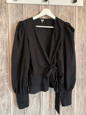 Svart ny omlottblus med puffärm NLY Trend 36, storlek S - Stilren svart omlottblus med långa puffärmar och breda manschetter. Blusen har knytband i midjan och en djup v-ringning framtill. Perfekt för dig som gillar en trendig och elegant look.